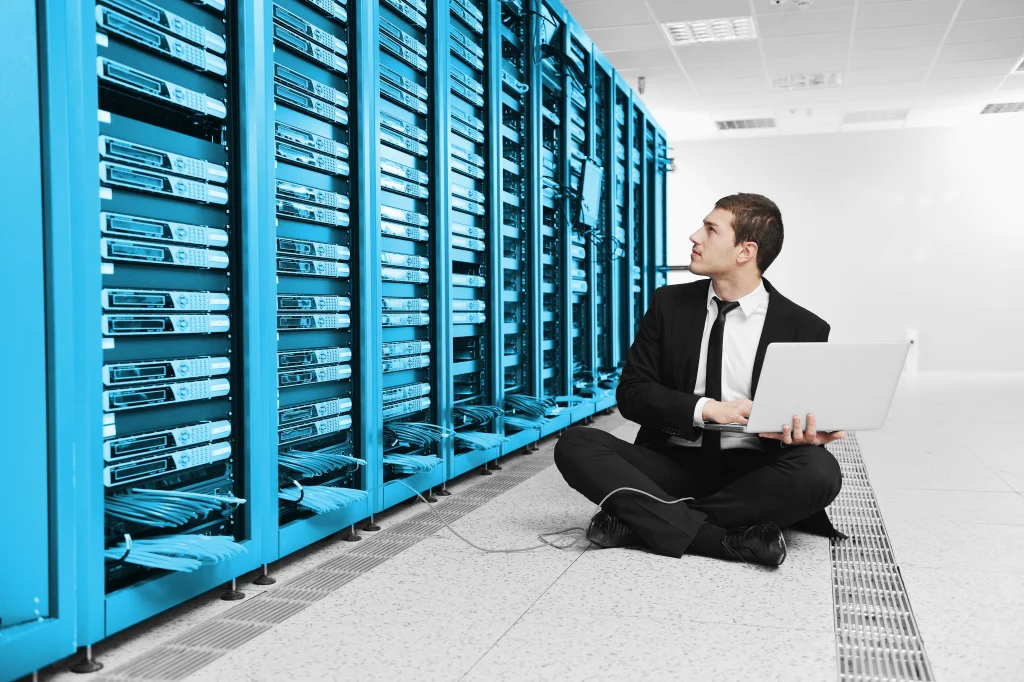 Ein IT-Experte sitzt entspannt auf dem Rechenzentrumsboden, vertieft in die Optimierung komplexer Netzwerkstrukturen auf seinem Laptop. Dieses Bild veranschaulicht die Kompetenz von CTE in der Bereitstellung hochverf&uuml;gbarer, zukunftssicherer und sicherheitsorientierter Netzwerkl&ouml;sungen, essentiell f&uuml;r die Leistungssteigerung in der vernetzten Fertigungsindustrie und die Realisierung der Industrie 4.0 Vision.